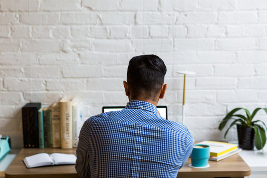 Image of man working at laptop, taken from behind. Man in blue shirt. White brick wall.